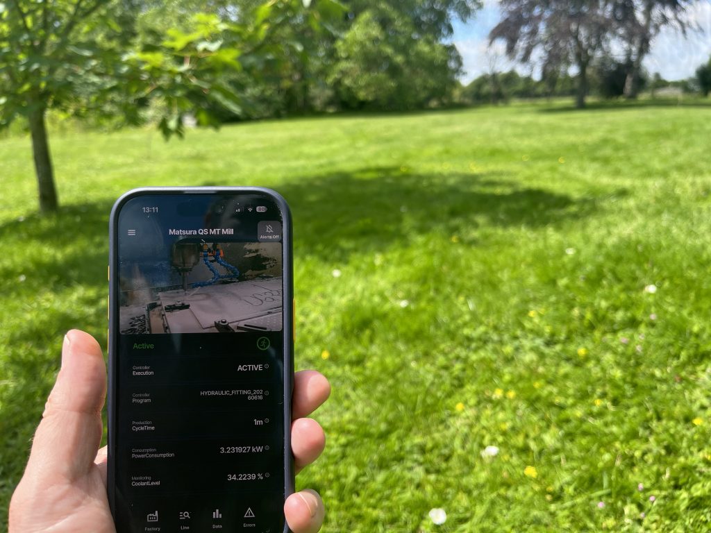 A smartphone running the RoboVigil app shows a live feed and status of a Matsura CNC mill, held in a sunlit park away from the factory.